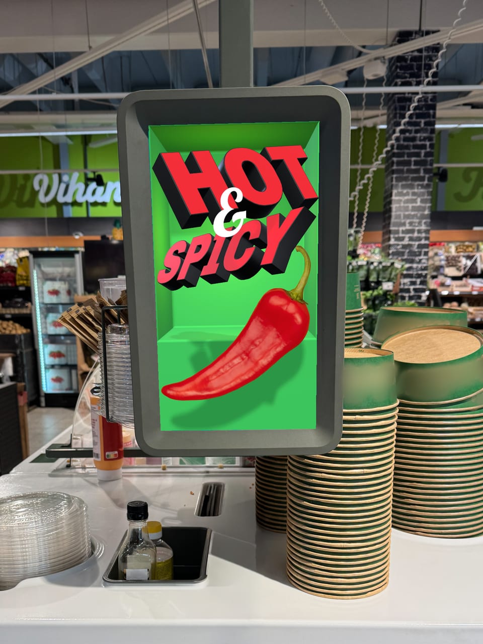 A small display on a supermarket deli counter. A red chilli lies in a green recess sunk into the screen, its tip crossing out over the frame, under the words HOT & SPICY.
