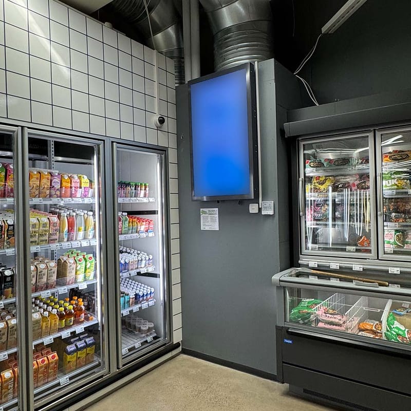A tall display mounted on a pillar in a shop, between the drinks fridges and the freezers, showing a plain blue screen.